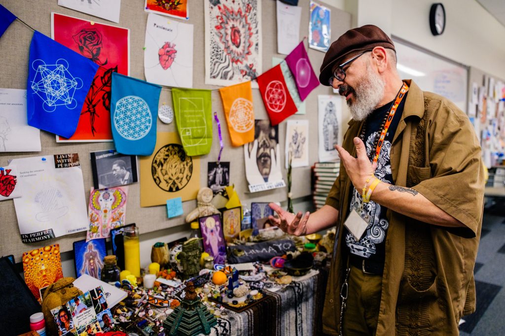 A man wearing a brown shirt and hat gestures while standing beside a colorful altar filled with various objects, with vibrant flags, artwork, and drawings displayed on the wall behind him.