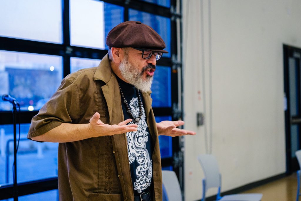 An older man with a gray beard, glasses, and a brown cap gestures expressively with his hands raised while speaking indoors near large windows and empty chairs.