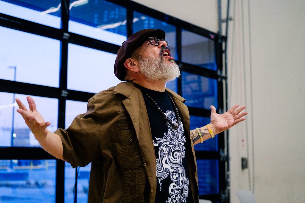 A bearded person wearing glasses, a brown shirt, and a hat stands indoors with arms raised and mouth open, appearing to speak or sing passionately. A large window is in the background.