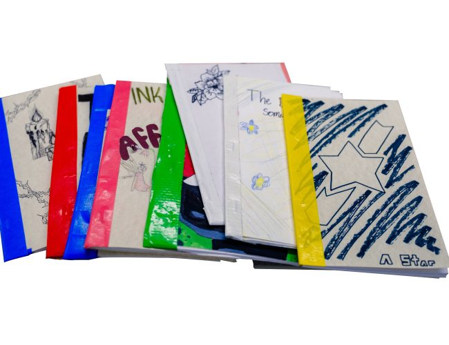 A stack of handmade, colorful booklets with illustrated and decorated covers, featuring drawings, text, and various colors of tape binding the spines, arranged on a white background.