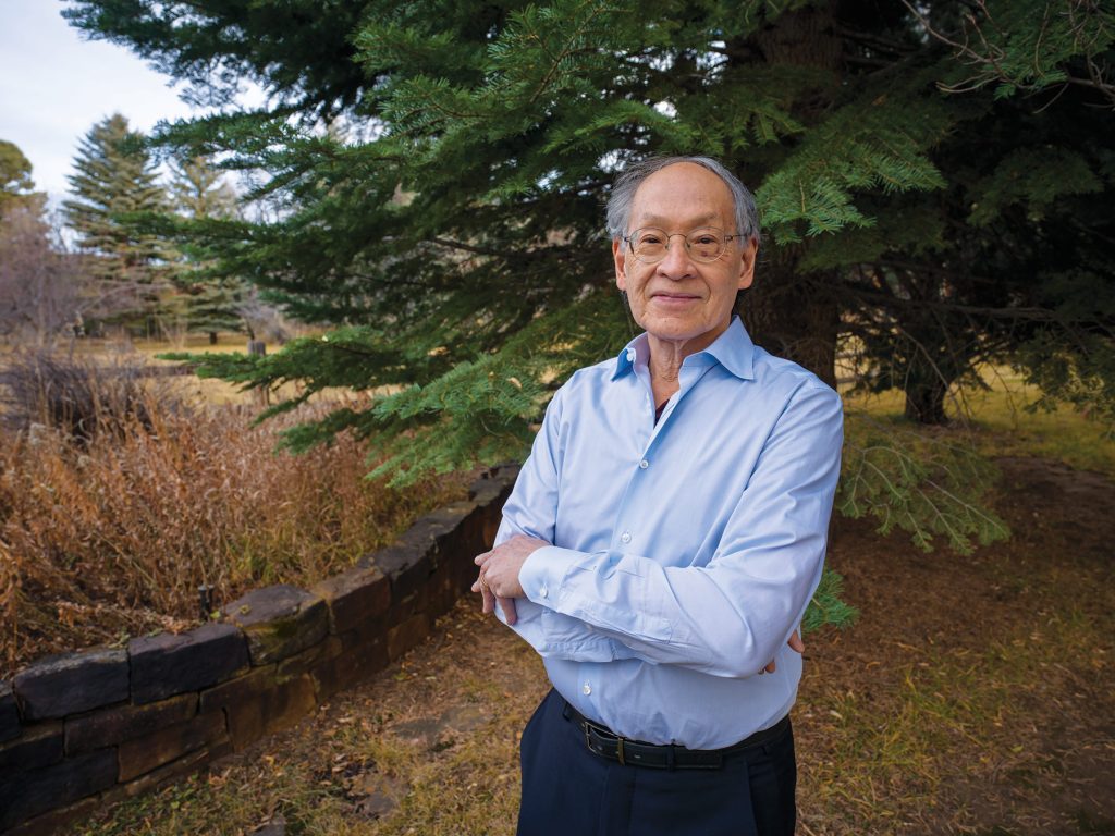 An older man wearing glasses and a light blue shirt stands outdoors with his arms crossed, smiling in front of a large pine tree and natural landscape.