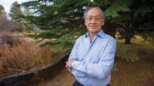 An older man wearing glasses and a light blue shirt stands outdoors with his arms crossed, smiling in front of a large pine tree and natural landscape.