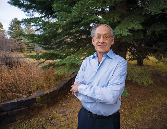 An older man wearing glasses and a light blue shirt stands outdoors with his arms crossed, smiling in front of a large pine tree and natural landscape.