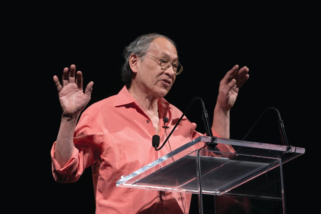 An older man with gray hair and glasses, wearing a coral shirt, speaks at a clear podium with microphones, gesturing with raised hands against a black background.