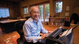 An older man in a light blue shirt smiles while sitting at a desk and typing on a typewriter in a cozy, well-lit home office filled with bookshelves and wooden furniture.