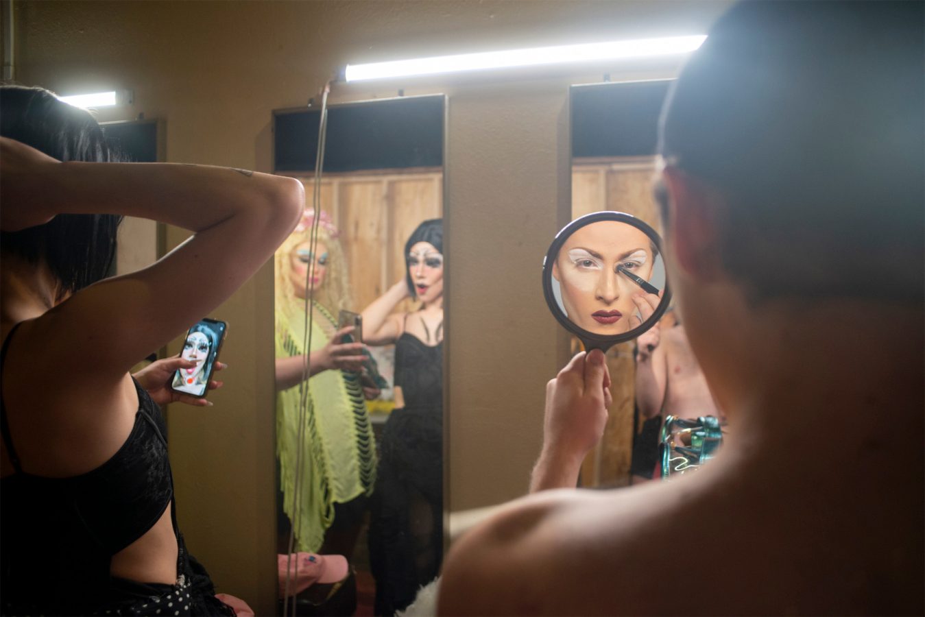 A person applies makeup using a handheld mirror, while two others, one in costume, look on and react in the background; another holds a phone displaying a reference image.
