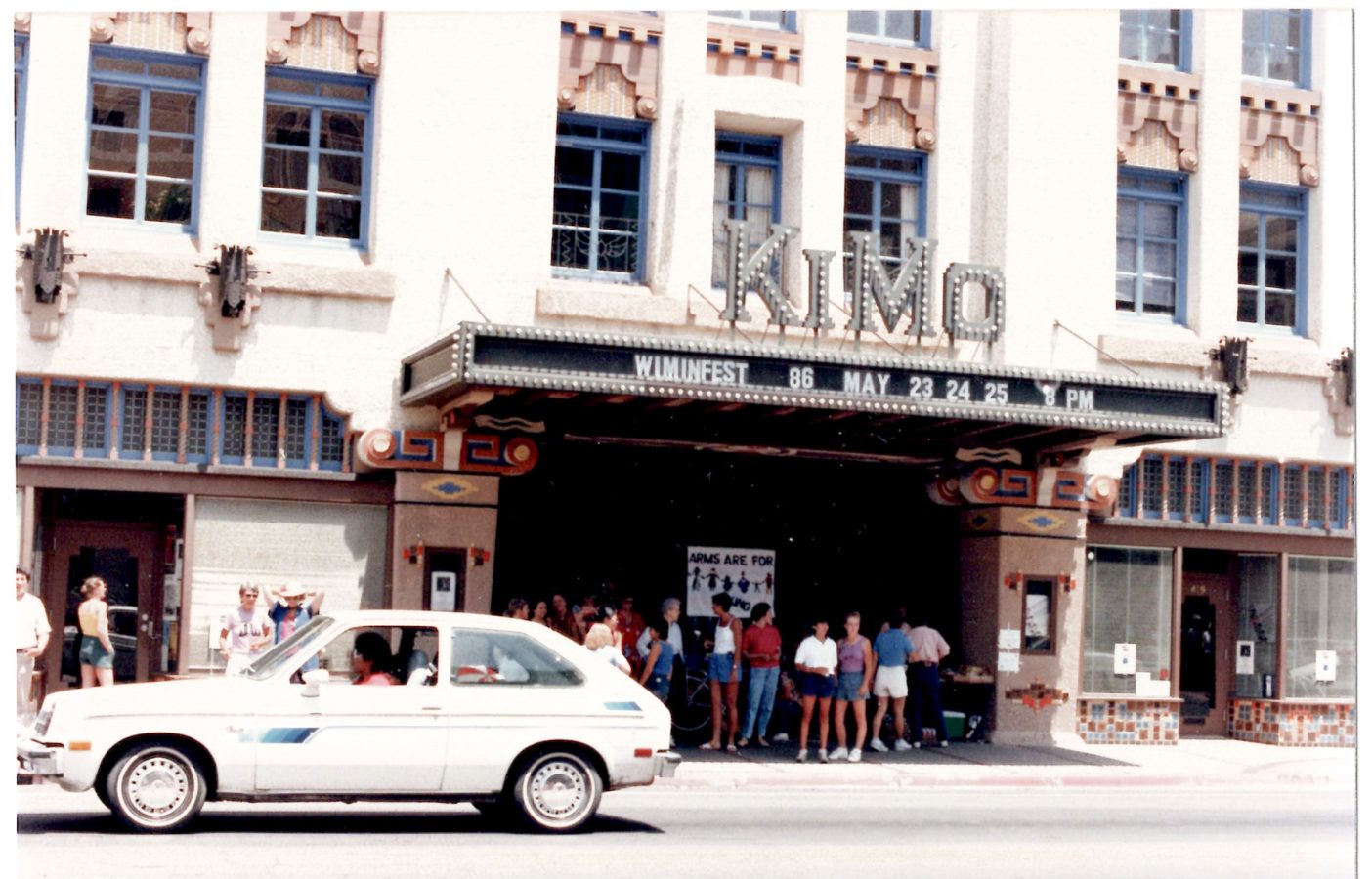 A white car is parked in front of the KiMo Theatre, where a crowd stands under the marquee advertising “WIMMFEST 86 MAY 23 24 25.”.