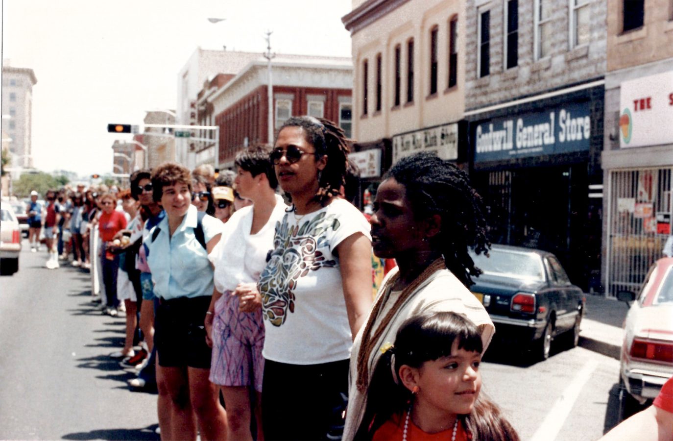 A diverse group of people stand holding hands across a city street in front of storefronts, forming a human chain during a public demonstration.