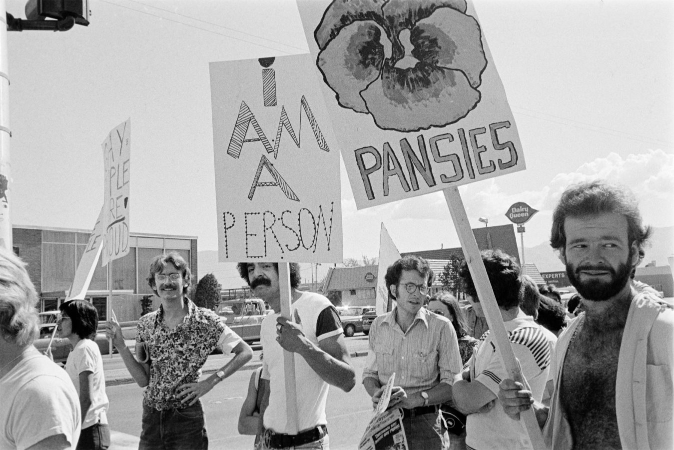 Black and white photo of people standing on a street holding protest signs, including one that says “I am a person” and another that says “PANSIES” with a drawing of a flower.