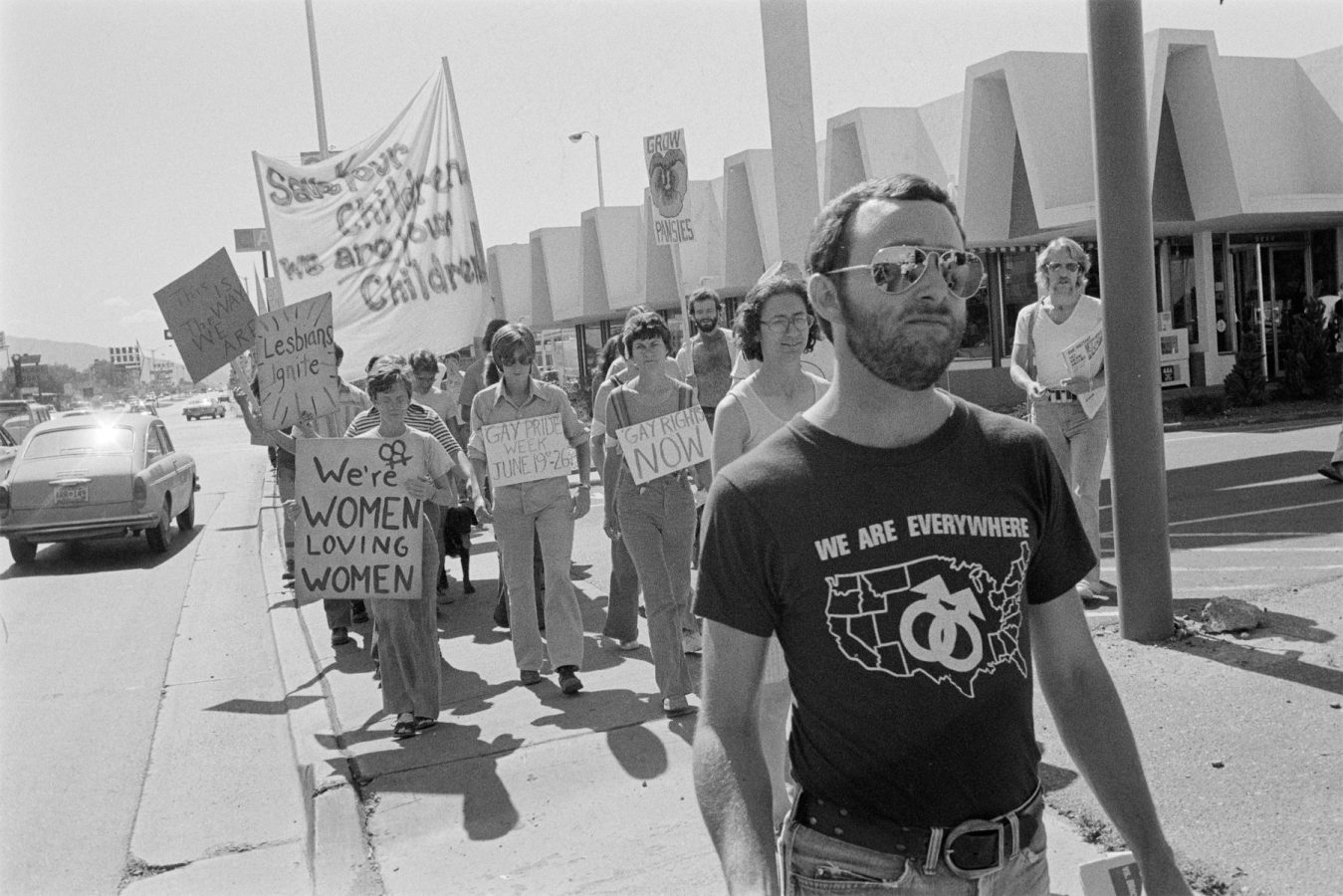 A group of people march on a sidewalk holding protest signs with LGBTQ+ messages; a man in front wears a “We Are Everywhere” shirt.