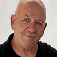 A bald older man with a mustache, wearing a black polo shirt, looks at the camera against a plain white background.