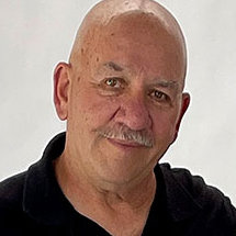 Bald older man with a mustache wearing a black collared shirt, posing in front of a plain white background.