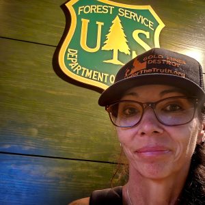 A woman wearing glasses and a cap with an environmental message stands in front of a U.S. Forest Service Department of Agriculture sign.
