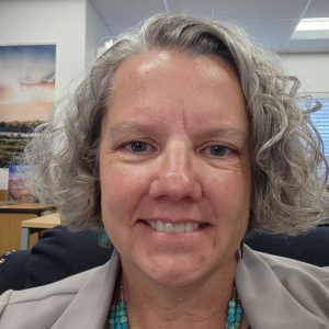 A middle-aged woman with short curly gray hair smiles at the camera in an indoor office setting.