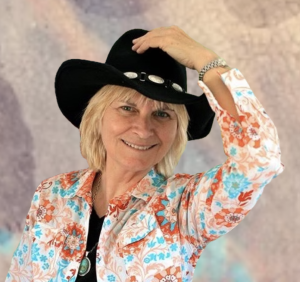 A smiling woman wearing a black cowboy hat and a colorful floral shirt touches the brim of her hat, posing in front of a blurred background.