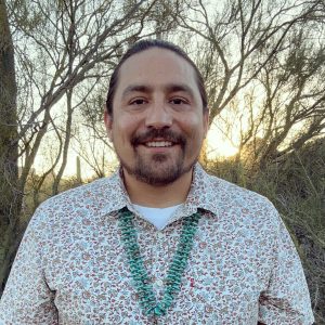 A man with long hair pulled back, wearing a patterned shirt and turquoise beaded necklace, stands outdoors with trees and the sun setting in the background.