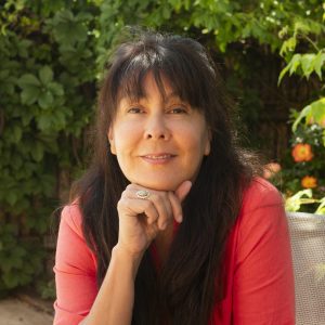 A woman with long dark hair wearing a coral shirt sits outdoors, resting her chin on her hand, with greenery and flowers in the background.