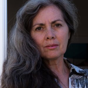 A woman with long gray hair and a patterned shirt looks directly at the camera with a neutral expression.