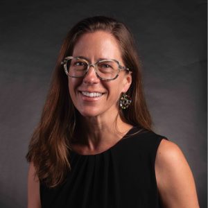 Woman with long brown hair, wearing glasses, a sleeveless black top, and colorful earrings, smiles against a dark gray background.