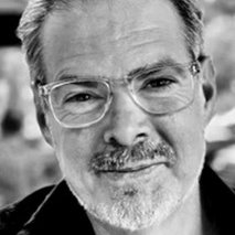 A middle-aged man with glasses and a short gray beard looks at the camera, wearing a dark shirt in a close-up black and white photo.