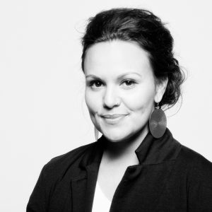 Black and white portrait of a woman with dark hair pulled back, wearing large round earrings, a dark jacket, and a slight smile, against a plain light background.