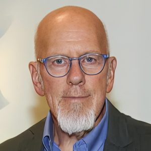 A bald man with a white goatee and blue-rimmed glasses wearing a dark jacket and blue shirt stands against a plain light background.