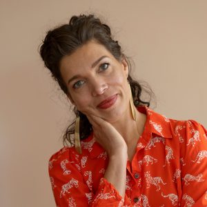 A woman with wavy brown hair wearing a red patterned shirt and gold earrings, resting her head on her hand, stands against a beige background.