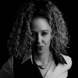 Black and white portrait of a woman with curly hair wearing a collared shirt and a necklace, looking at the camera against a black background.
