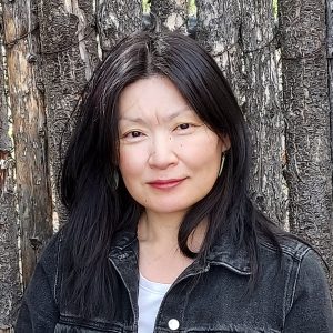 A woman with straight black hair wearing a black denim jacket stands in front of tree trunks, looking at the camera with a neutral expression.