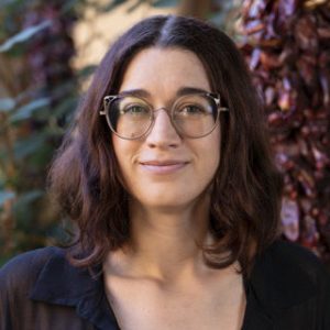 A person with shoulder-length brown hair and glasses is standing outdoors, wearing a black top, with plants and red chili peppers visible in the background.