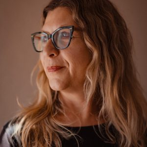 A woman with long wavy blonde hair and glasses looks to the side, wearing a black top, against a neutral background.