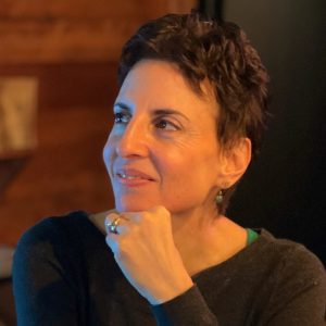 A person with short dark hair, wearing a dark top, rests their chin on their hand and looks slightly to the side with a thoughtful expression.
