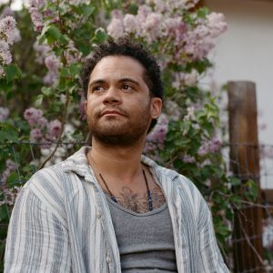 A man with short curly hair and a tattoo on his chest wears a striped shirt and sits in front of blooming lilac bushes, looking off to the side.