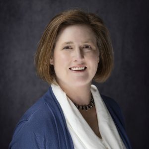 A woman with short brown hair wears a blue cardigan, white blouse, and black beaded necklace, smiling in front of a dark gray background.