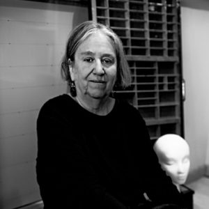 An older person with gray hair sits indoors next to a mannequin head, with shelves visible in the background.