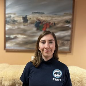 A woman wearing a navy STAFF t-shirt stands indoors in front of a fuzzy beige blanket and a framed landscape painting.