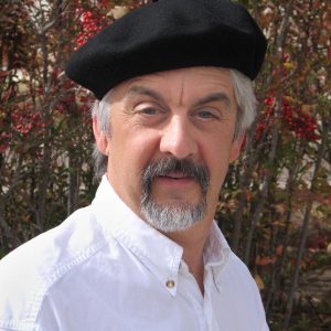 Middle-aged man with gray hair and mustache wearing a black beret and white shirt, standing outdoors in front of berry bushes.