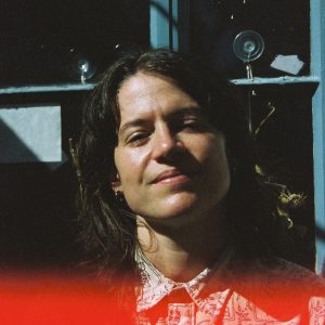 A person with shoulder-length brown hair and a patterned shirt stands in sunlight, with a faint red blur at the bottom of the image and a window in the background.