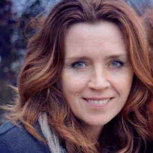 A woman with long, wavy brown hair and blue eyes is smiling at the camera outdoors.