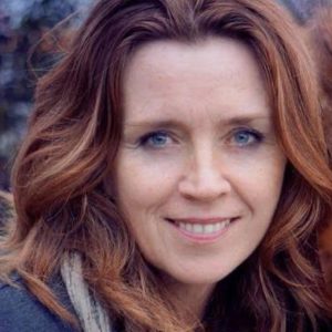 Woman with wavy, shoulder-length auburn hair, wearing a grey top and scarf, smiling outdoors with blurred foliage in the background.