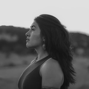 Woman with long hair and earrings standing outdoors, shown in profile, looking into the distance. The image is in black and white.