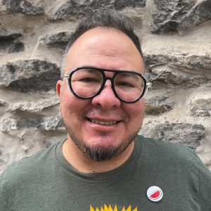 A person with short hair, glasses, and a goatee smiles in front of a stone wall, wearing a green shirt and a watermelon pin.