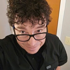 Person with curly dark hair and glasses wearing a black shirt, smiling and looking up at the camera indoors near a door and light switch.